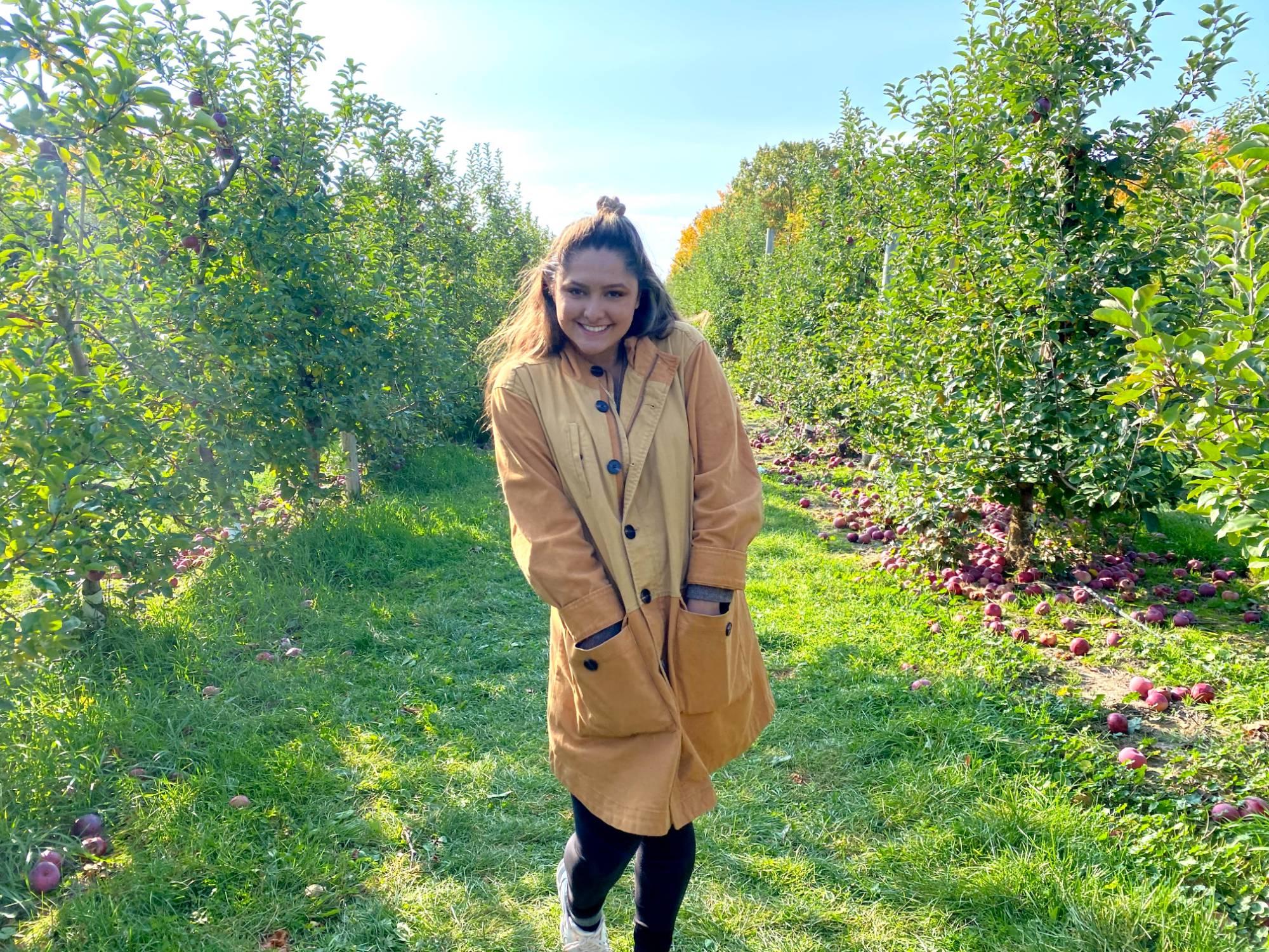 girl with brown hair and a tan coat standing in an apple orchard